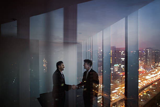 Levně Plakát, Obraz - Businessmen shaking hands in office at night, Shannon Fagan, 40 × 26.7 cm