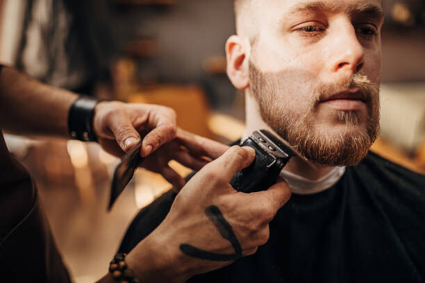 Levně Plakát, Obraz - Male barber cutting beard of client in barber shop, South_agency, 40 × 26.7 cm